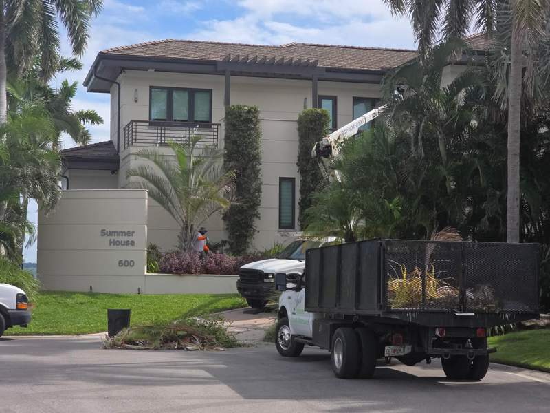 Bucket truck trimming palms at a large two-story home while a crew member rakes up fronds beside a loaded dump truck.